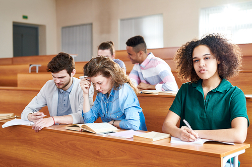 Content confident African-American student girl with curly hair sitting at university desk and making notes in workbook while  in lecture room, other students reading books