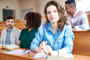 Smiling attractive young female student with curly hair sitting at university desk with workbook and pen and  in university lecture room