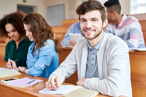 Smiling positive handsome young male student with beard sitting at wooden desk together with groupmates and  in university
