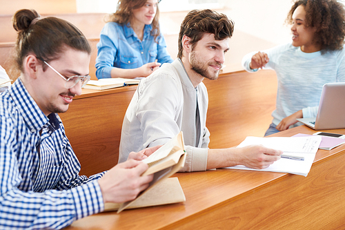 Positive curious handsome bearded student talking to lecturer while participating in discussion at university class, multiethnic students sitting at table in row