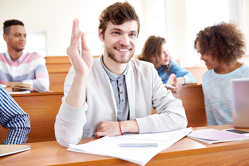 Cheerful excited handsome bearded student boy sitting at wooden desk with open workbook and raising hand while having question to lecturer