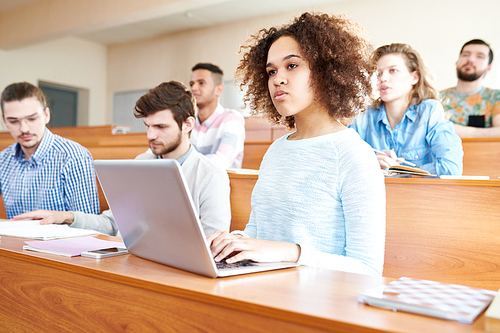 Pensive concentrated young multiethnic students sitting at university desk and attentively listening to lecturer, serious African-American girl with Afro hairstyle typing on laptop