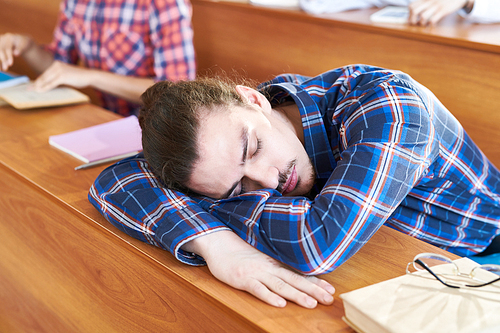 Tired handsome hipster student boy in checkered shirt lying on wooden desk and sleeping during lecture in university
