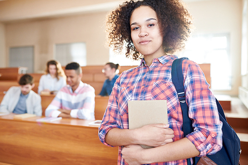 Smiling content attractive African-American student girl with curly hair wearing satchel on shoulder holding small textbook and  in university room