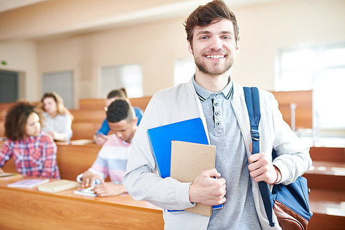 Cheerful positive ambitious handsome young guy in casual jacket smiling at camera and holding satchel and workbooks in lecture room