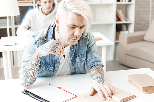 Hipster-like Caucasian man with tattoos and dyed blond hair sitting at desk in college library and reading book carefully