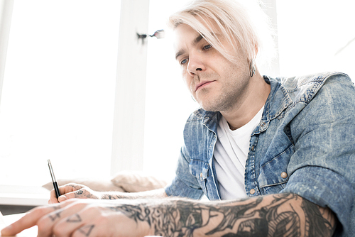 Low angle view of hipster-like tattooed man with dyed blond hair and earrings sitting at desk and reading book thoroughly