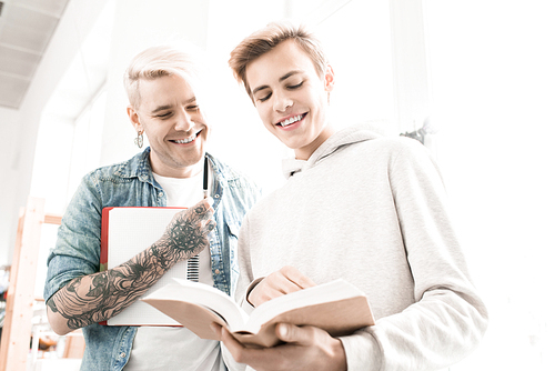 Hipster-like tattooed adult student and his young college mate looking at textbook and smiling happily, low angle view