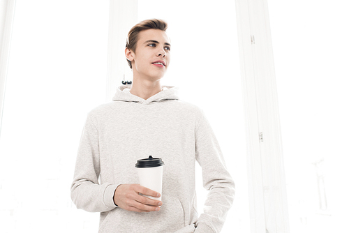 Handsome male teenager in white hoodie standing against window with coffee to-go, looking aside and smiling, low angle view