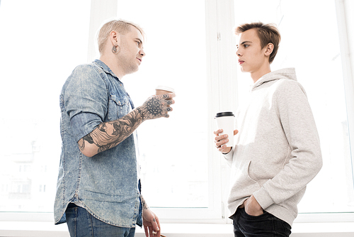 Low angle view of hipster-like man with tattooed hands talking to teenager while standing against window with takeaway coffee