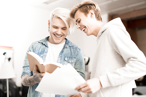 Caucasian hipster-like tattooed male student and his friend looking at pictures in textbook and smiling joyfully