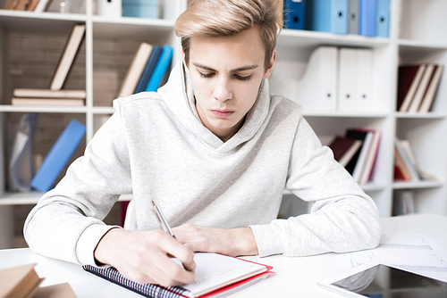 Young concentrated Caucasian male student writing notes in copybook while sitting at desk in classroom