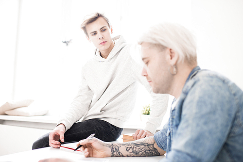 Young Caucasian male student sitting on desk and listening attentively to his adult college mate with tattooed hands