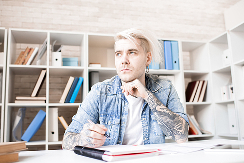 Caucasian adult man with tattoos, earrings and dyed hair sitting at desk in study room and looking away thoughtfully