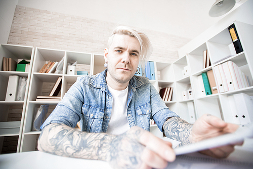 Low angle view of Caucasian adult student with tattoos, dyed hair and earrings sitting in study room and 