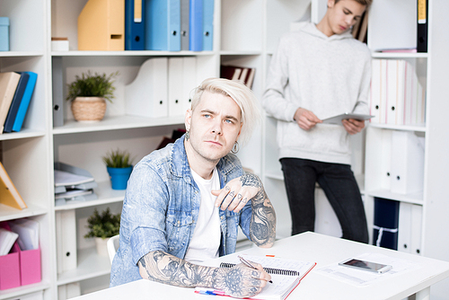 Caucasian adult student with tattoos and dyed hair looking aside pensively while preparing for exam in study room