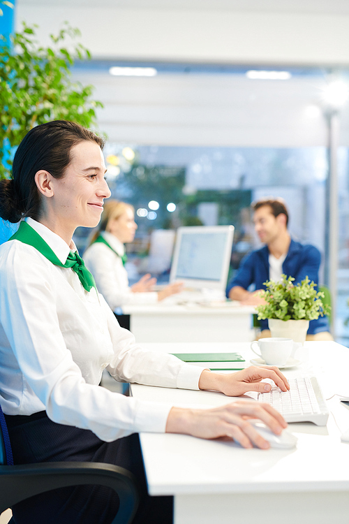 Smiling satisfied female bank employee in white blouse and green neck scarf sitting at table and using computer while preparing report in modern office
