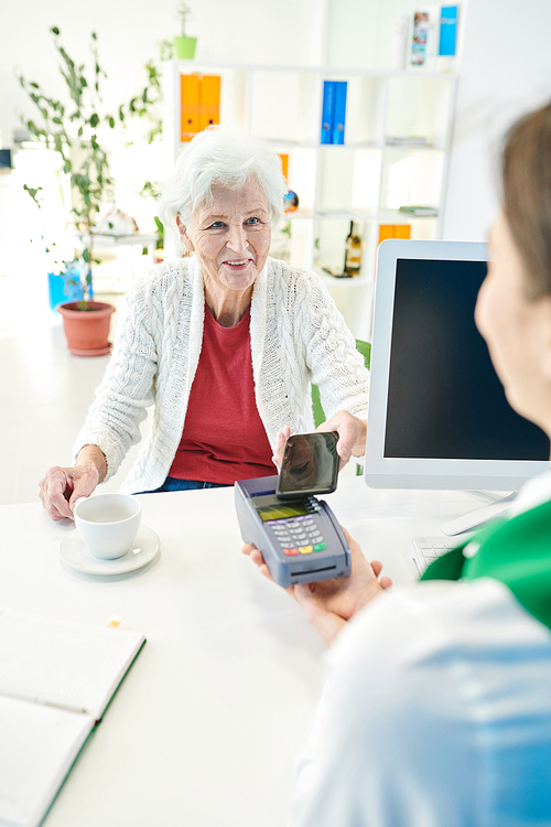 Cheerful attractive senior female bank client in casual cardigan sitting at table and drinking coffee while paying with smartphone for bank service