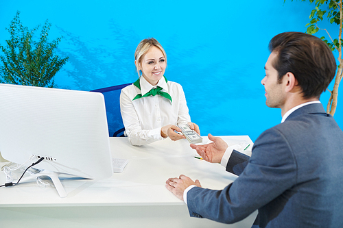 Cheerful confident attractive bank manager in green scarf on neck sitting at table with computer and giving money from deposit account to businessman