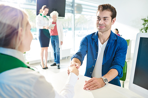 Content satisfied handsome young man in casual shirt sitting at table and making handshake with bank sales representative while concluding deal with bank