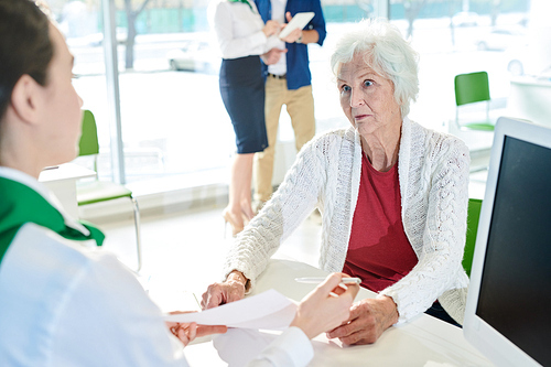 Shocked senior lady customer in white cardigan sitting at table and looking with confusion at bank specialist while listening to contract explanations in office