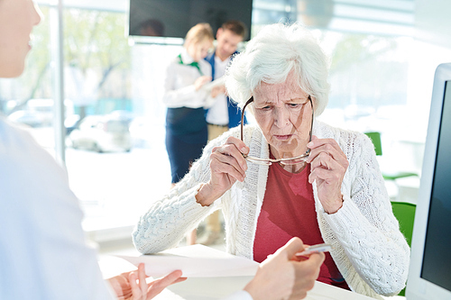 Serious confused senior lady with gray hair sitting at table and looking through eyeglasses while examining document shown by manager in bank office