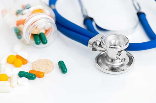 High angle close up of assorted colorful medication drugs and vitamins spilling from bottle over white background next to stethoscope on table in doctors office