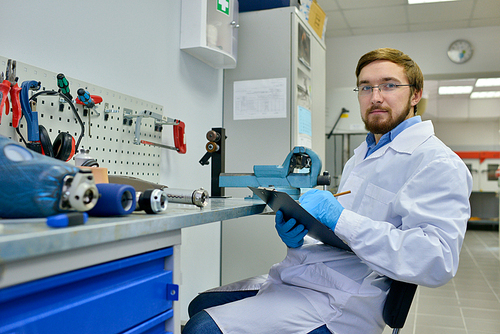 Portrait of young medical engineer posing in laboratory sitting by table with orthopedic equipment and  holding clipboard