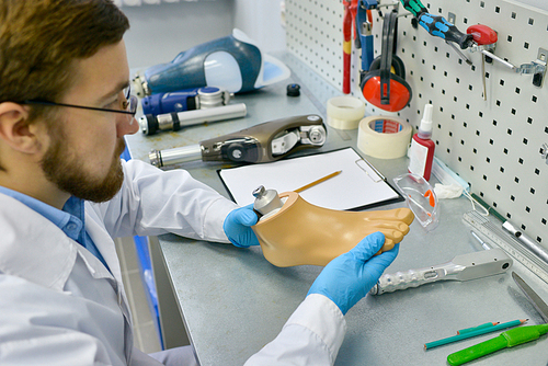 Side view portrait of young doctor holding artificial foot at desk in office, checking it for quality and making adjustments