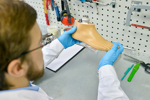 High angle portrait of young doctor holding artificial foot at desk in office, checking it for quality and making adjustments