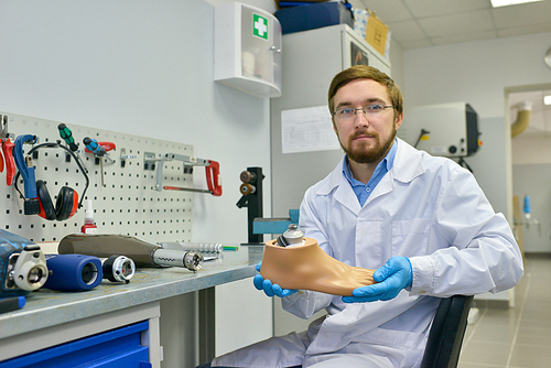 Portrait of young medical engineer posing in laboratory sitting by table with orthopedic equipment and  holding artificial foot