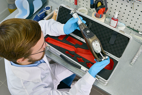 High angle view of  medical engineer holding prosthetic leg part at desk in office, checking it for quality and making adjustments