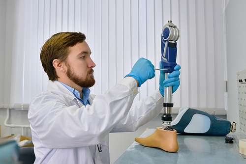 Side view of  prosthetics technician repairing prosthetic leg part at desk in office, checking it for quality and making adjustments