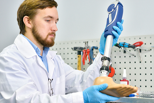 Portrait of young  prosthetist holding prosthetic leg  checking it for quality and making adjustments while working in modern laboratory, copy space