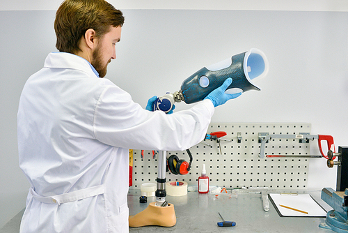 Back view portrait of young  prosthetics technician holding prosthetic leg  checking it for quality and making adjustments while working in modern laboratory, copy space