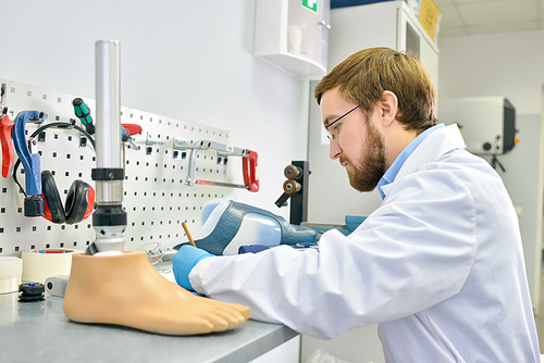 Side view portrait of  young prosthetics technician  making prosthetic leg at desk in office and writing notes, copy space