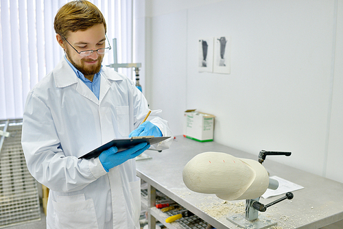 Portrait of young prosthetics technician  writing notes on clipboard while checking prosthesis molds working in  modern design laboratory, copy space