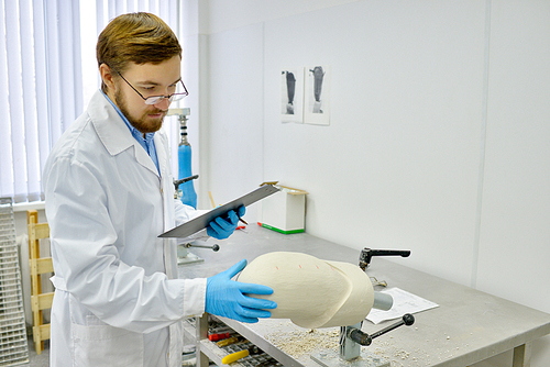 Portrait of young prosthetics technician  checking prosthesis molds and holding clipboard while working in  modern design laboratory, copy space