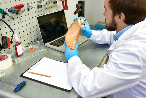 Portrait of young  prosthetist holding prosthetic leg  checking it for quality and making adjustments while working in modern laboratory, copy space