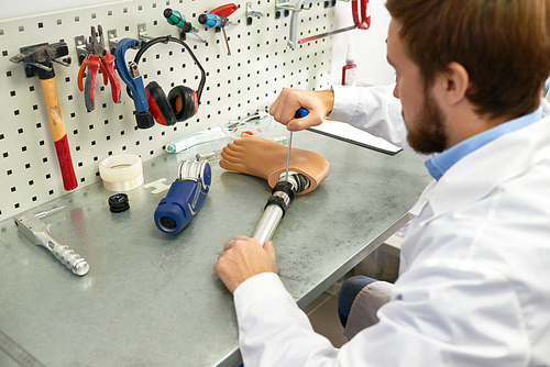 Portrait of  prosthetist at work, over the shoulder view of young man assembling artificial foot sitting at table in modern laboratory