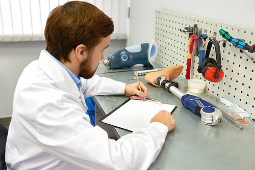 Portrait of  bearded prosthetics technician  making prosthetic leg at desk in office and writing notes on clipboard, copy space