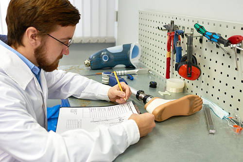 Side view portrait of  bearded prosthetics technician  making prosthetic leg at desk in office and writing notes on clipboard, copy space