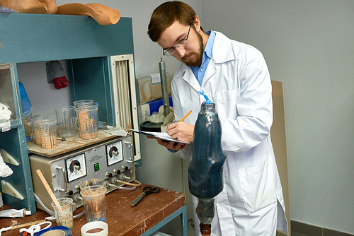 Portrait of young  prosthetist working in laboratory writing notes on clipboard while examining prosthetic leg molds