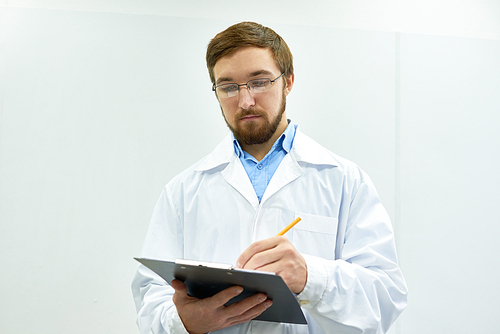 Portrait of young bearded doctor wearing glasses standing against white wall writing on clipboard, copy space