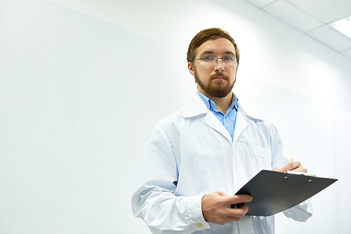 Low angle  portrait of  bearded doctor wearing glasses standing against white wall writing on clipboard and , copy space