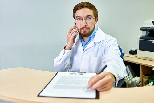 Portrait of friendly bearded doctor handing pen and contract to patient siting at desk in office and using smartphone