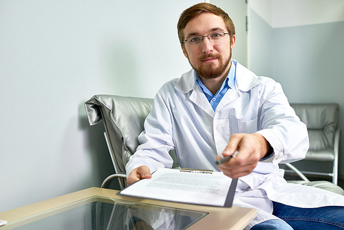 Portrait of friendly bearded doctor handing pen and form to patient siting at desk in office