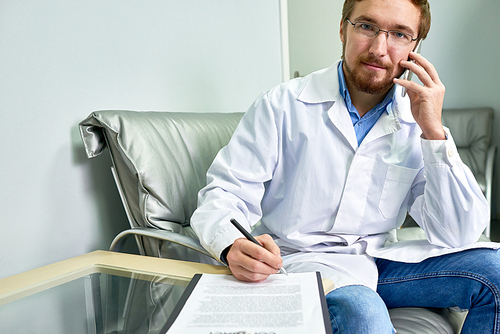 Portrait of friendly bearded doctor speaking by phone and  while signing contract form