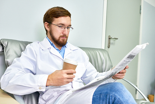 Portrait of bearded doctor relaxing in chair and reading newspaper during coffee break at work