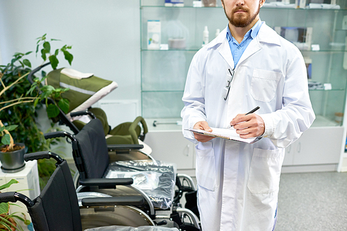 Mid-section of bearded doctor posing with clipboard in hospital hall next to row of wheelchairs, copy space